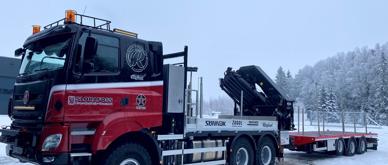 A red and black TATRA truck is parked on a snowy road. The truck has a large crane on the back and a flatbed trailer attached to it, in front of a snowy tree-covered mountain.