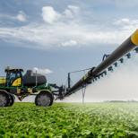 An agriculture sprayer, painted green and yellow, is applying pesticides to a field of soybeans.