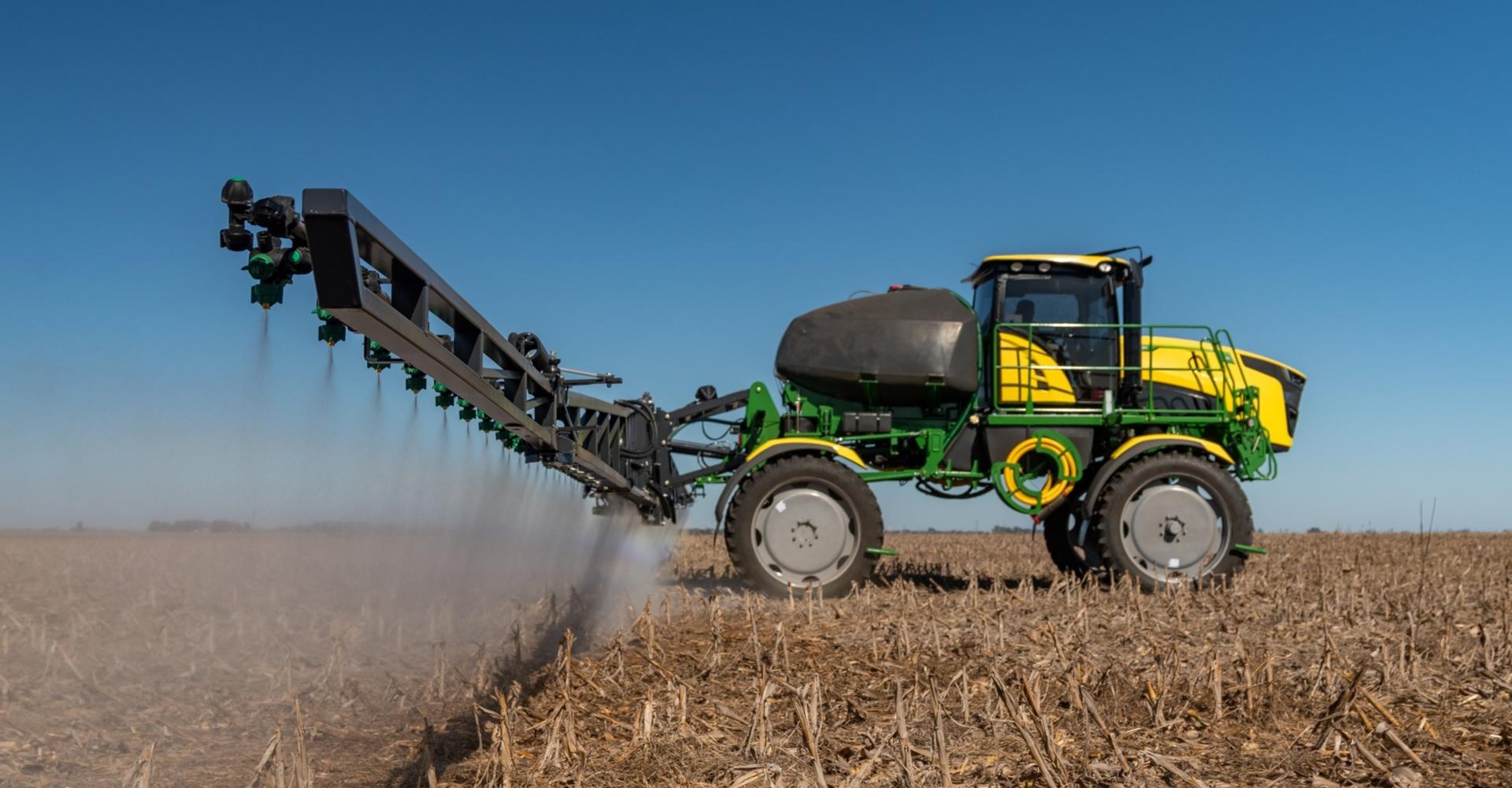 A yellow and green ag sprayer is spraying pesticides on a dry, brown field. The sprayer has a long boom with nozzles that are spraying the pesticides.